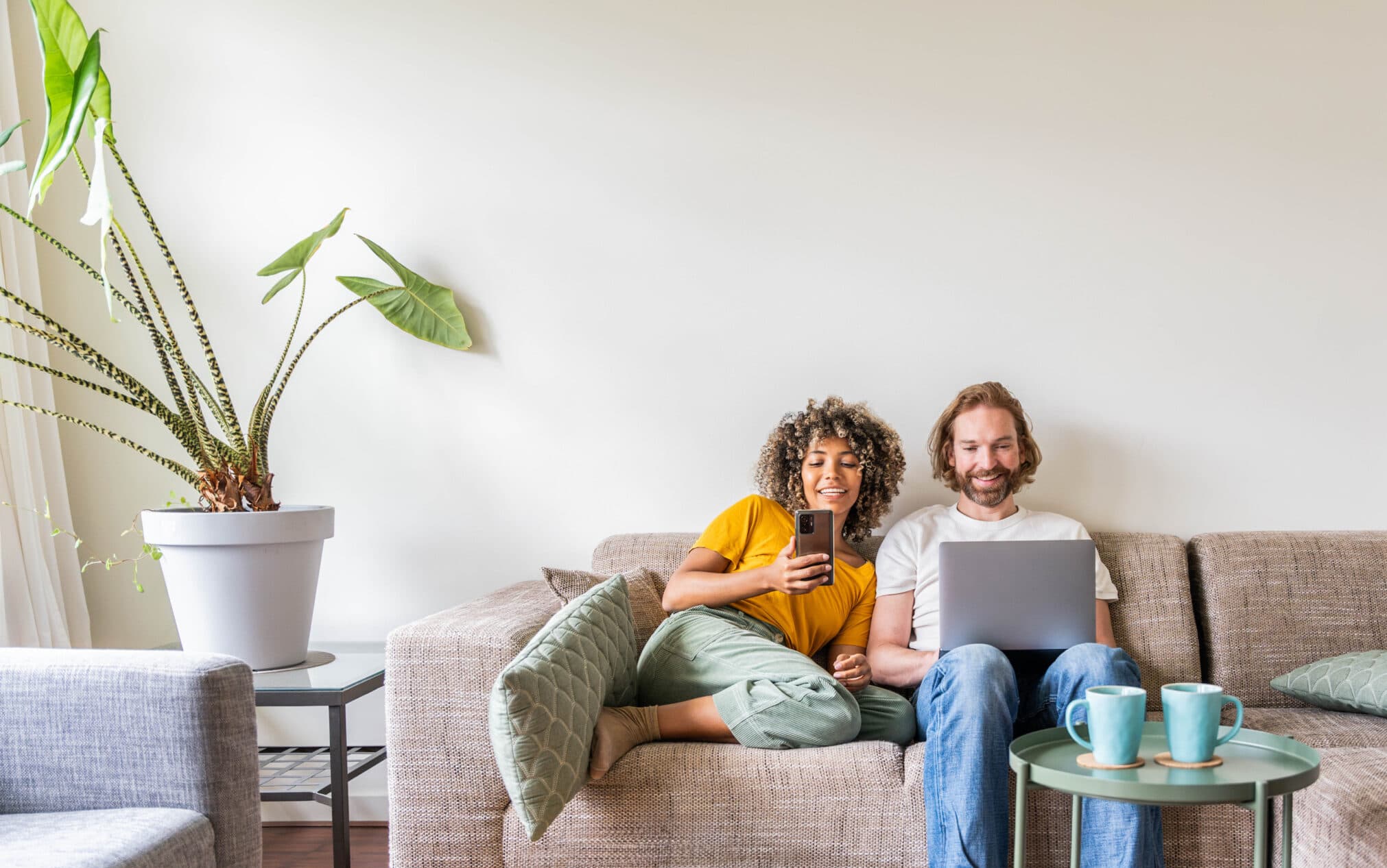Deux personnes assises sur un canapé moderne, utilisant un ordinateur portable et un smartphone, avec une table basse et des tasses, illustrant la connectivité et le confort des solutions digitales.