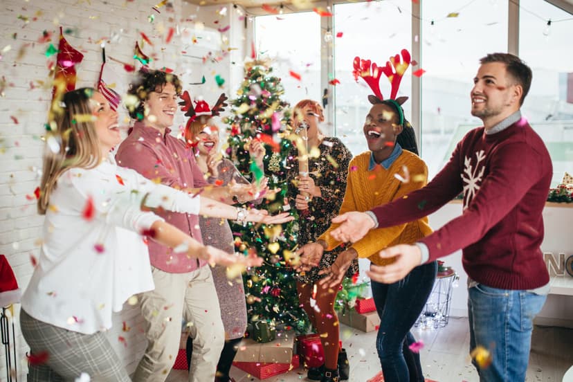 Groupe de personnes célébrant Noël dans un bureau, avec des décorations festives, un sapin de Noël et des confettis en l’air.